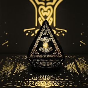 May include: A black, pyramid-shaped decorative lamp with intricate cut-out patterns. The lamp features a glowing light inside, casting shadows of Celtic knot designs and a Thor's hammer symbol onto the surrounding surface. The lamp is a unique home decor item.
