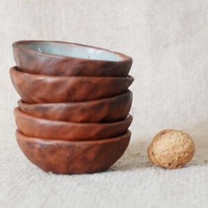 May include: A stack of six small, brown ceramic bowls with a light blue glaze on the top bowl. The bowls are stacked on a light brown fabric surface. A single brown walnut is in the foreground.