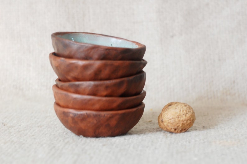 May include: A stack of six small, brown ceramic bowls with a light blue glaze on the top bowl. The bowls are stacked on a light brown fabric surface. A single brown walnut is in the foreground.