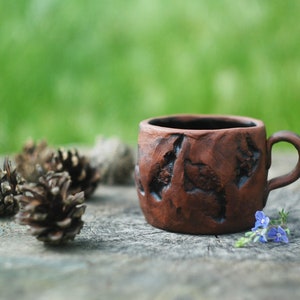 May include: A rustic brown ceramic mug with a textured surface sits on a wooden surface with pine cones and blue flowers.