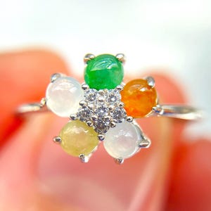 May include: A silver ring featuring a flower design with six colorful gemstones. The gemstones are green, orange, yellow, and white, with a cluster of small clear stones at the center. The ring is held in a person's hand.