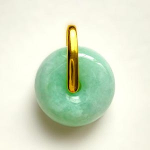 Puede incluir: Un colgante redondo de jade verde claro con una fianza dorada. El jade tiene una superficie lisa y pulida y un agujero en el centro. La fianza dorada tiene una forma de barra vertical simple. El colgante está sobre un fondo blanco.