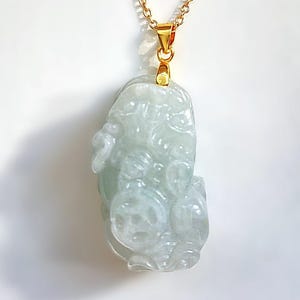 Könnte beinhalten: Eine goldfarbene Halskette mit einem hellgrünen Jade-Anhänger. Der Anhänger ist mit komplizierten Details geschnitzt und hängt an einer kleinen goldenen Öse. Die Halskette wird vor einem weißen Hintergrund präsentiert.
