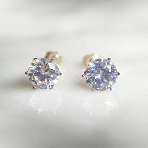 May include: A pair of clear, round-cut stud earrings. Each earring has a large, faceted gemstone set in a silver-coloured metal. The earrings are displayed on a white, reflective surface.