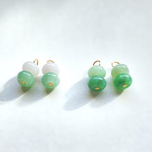 May include: Earrings featuring a gold hook and two jade beads in shades of green and white. The jade beads are smooth and round, with a small gold accent on the bottom. The earrings are displayed on a white background.