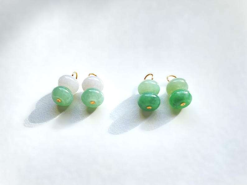 May include: Earrings featuring a gold hook and two jade beads in shades of green and white. The jade beads are smooth and round, with a small gold accent on the bottom. The earrings are displayed on a white background.
