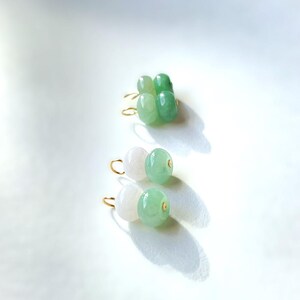 May include: Close-up of three pairs of earrings. The earrings feature gold-colored hooks and are adorned with jade beads in shades of white and green. The earrings are displayed on a white surface, with shadows adding depth.
