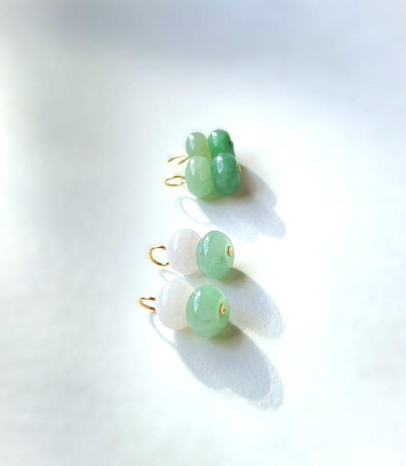 May include: Close-up of three pairs of earrings. The earrings feature gold-colored hooks and are adorned with jade beads in shades of white and green. The earrings are displayed on a white surface, with shadows adding depth.