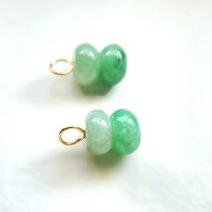 May include: Two jade pendants with a light green color and a gold-colored loop. Each pendant is made of two connected, rounded jade beads. The jade has a translucent quality, with variations in the green hue.