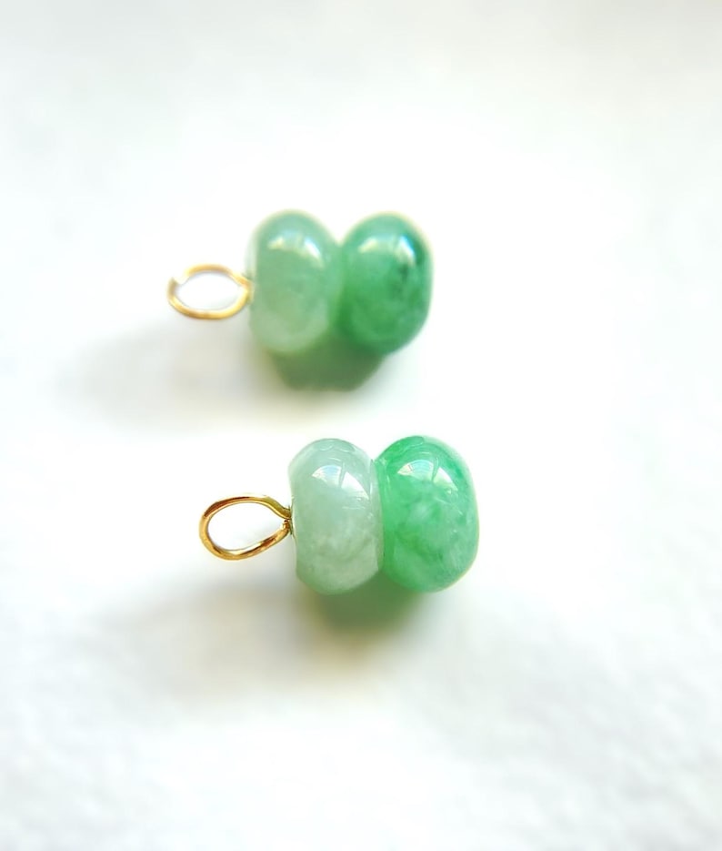 May include: Two jade pendants with a light green color and a gold-colored loop. Each pendant is made of two connected, rounded jade beads. The jade has a translucent quality, with variations in the green hue.