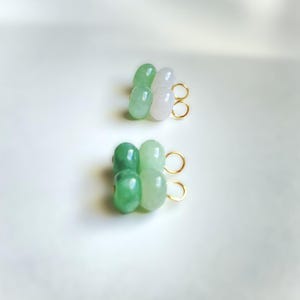May include: Two pairs of earrings, each featuring four jade beads in shades of green and white. The beads are strung together and attached to gold-colored hoops. The earrings are displayed on a white surface.