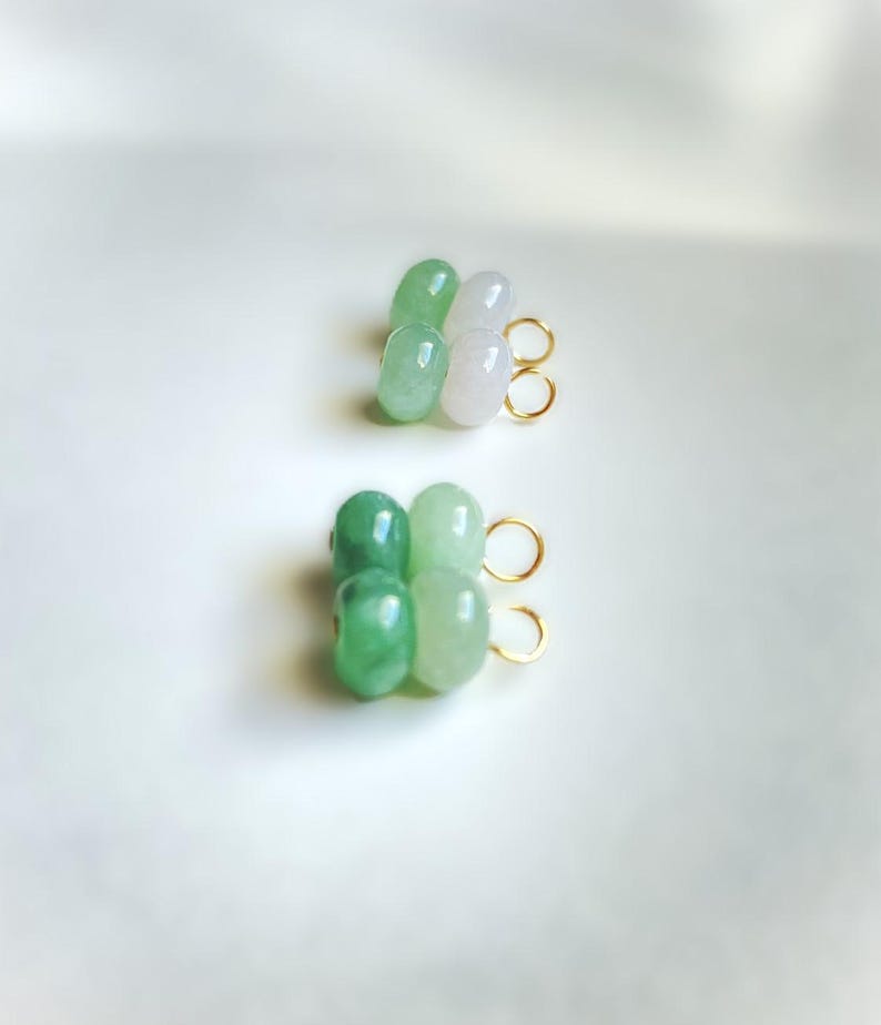 May include: Two pairs of earrings, each featuring four jade beads in shades of green and white. The beads are strung together and attached to gold-colored hoops. The earrings are displayed on a white surface.