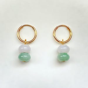 May include: A pair of gold hoop earrings, each featuring two jade beads: a light lavender top bead and a green bottom bead. The earrings are displayed against a white background.