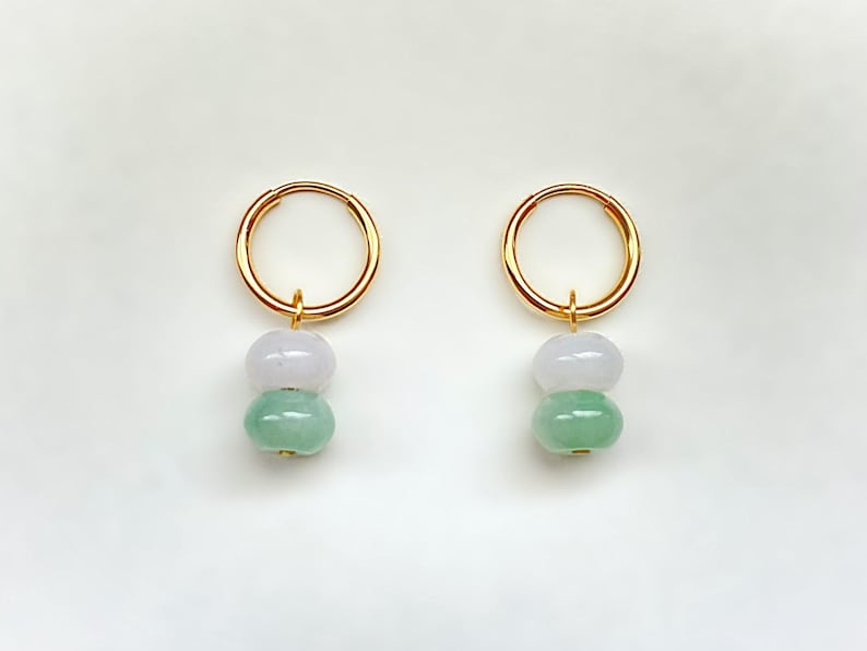 May include: A pair of gold hoop earrings, each featuring two jade beads: a light lavender top bead and a green bottom bead. The earrings are displayed against a white background.