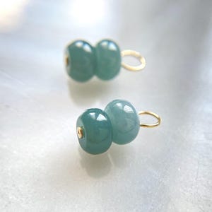 May include: Two light teal-colored, double-beaded earrings with gold-colored loops. The beads have a smooth, glossy finish and are connected. The earrings are set against a soft, blurred background.