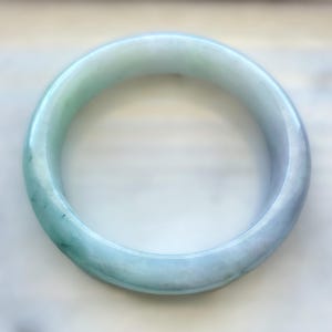 Op de afbeelding: Een gladde, cirkelvormige armband gemaakt van gepolijste steen. Het sieraad heeft een doorschijnend uiterlijk met een verloop van zachte mintgroene en licht lavendelkleurige tinten, met natuurlijke interne variaties en een glanzende afwerking.