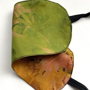 Peut inclure: Un masque de sommeil vert et doré avec un motif floral. Le masque est fait d'une matière soyeuse et possède une sangle élastique noire. L'intérieur du masque est de couleur dorée avec un motif floral rose.