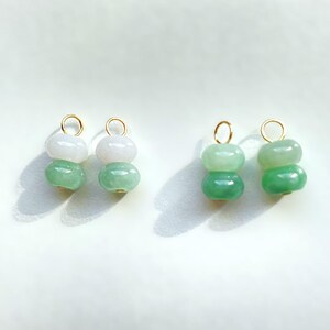 May include: Four jade gemstone charms. Each charm features two stacked, rounded beads in shades of green and white, with a gold-colored loop at the top. The charms are displayed on a white surface.