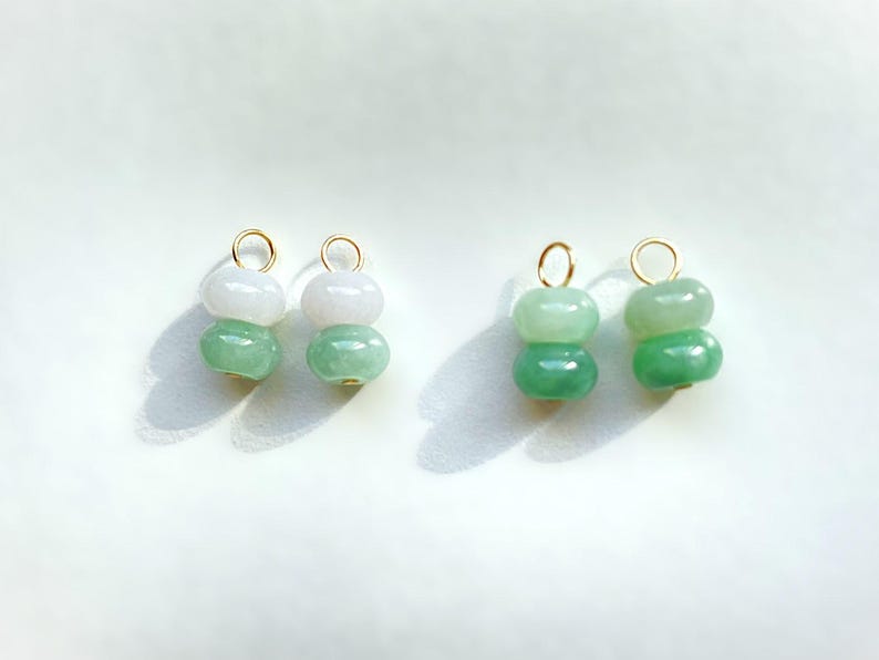 May include: Four jade gemstone charms. Each charm features two stacked, rounded beads in shades of green and white, with a gold-colored loop at the top. The charms are displayed on a white surface.