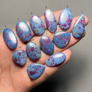 May include: A selection of twelve polished ruby in zoisite gemstones. The stones are in various shapes, including oval, teardrop, and irregular forms. The gemstones display a mix of vibrant red ruby inclusions within a blue-green zoisite matrix.