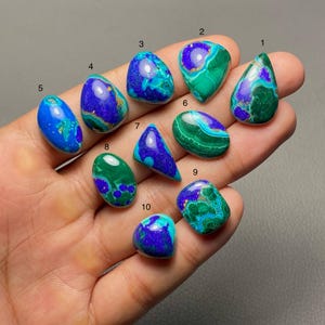 May include: A collection of ten polished gemstones in various shapes, held in a hand. The stones display a mix of deep blues and greens, with some featuring swirling patterns and others a more solid color. The gemstones are numbered 1 through 10.