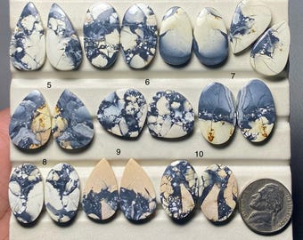 May include: A collection of ten polished gemstones in various shapes, including teardrop and oval. The stones feature a mix of white, gray, and blue patterns, with a nickel coin for size comparison. The gemstones are displayed on a white surface.