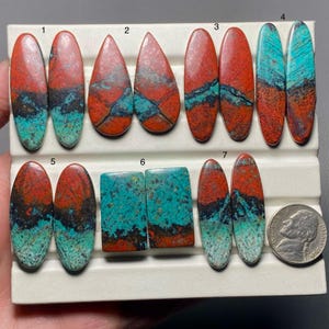 May include: A collection of polished gemstones in various shapes, including oval and teardrop. The stones display a striking combination of red, turquoise, and black colors. A US quarter coin is included for size reference. These stones are likely for jewelry making.