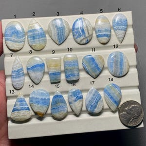 May include: A collection of 18 blue and white banded agate cabochons, arranged in rows on a white display board. The cabochons are various shapes and sizes, including oval, teardrop, and heart shapes.