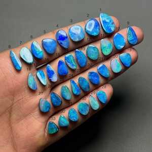 May include: A collection of 33 polished opal gemstones in various shapes and sizes, displayed on a hand. The opals exhibit a range of blue, green, and white colors, with visible internal patterns. Each stone is numbered for identification.