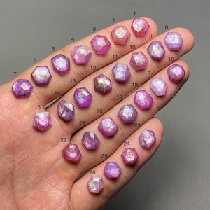 Puede incluir: Una colección de veintiséis gemas hexagonales facetadas, en varios tonos de rosa y púrpura. Las gemas se sostienen en una mano, mostrando sus superficies pulidas y variaciones de color únicas. Cada piedra está numerada para su identificación.
