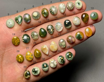 May include: A selection of 34 polished gemstones in diverse shapes and colours, including green, yellow, brown, and white. Each stone is numbered, displaying a variety of patterns and textures. The gemstones are held in a hand, against a neutral backdrop.