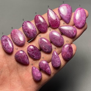 以下が含まれることがあります： ピンクと紫の色合いの、楕円形と涙滴型の研磨された宝石のコレクション。 宝石は手の中にあり、滑らかで反射する表面が示されています。