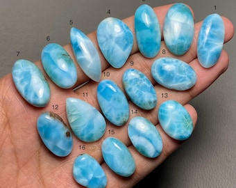 Puede incluir: Una colección de dieciséis piedras preciosas de larimar pulidas en varias formas, desde ovaladas hasta en forma de lágrima. Las piedras muestran tonos azules con vetas blancas, sostenidas en una mano.