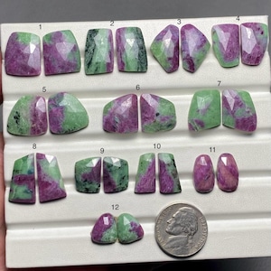 May include: Twelve pairs of green and purple gemstone cabochons, numbered 1 through 12, arranged in rows on a white surface. A US quarter is included for scale.