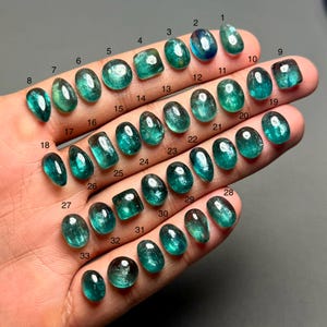May include: A hand holds 33 polished gemstones, each with a unique shape and size. The gems vary in colour from deep teal to light blue-green, some with internal patterns. Each gem is numbered for identification.