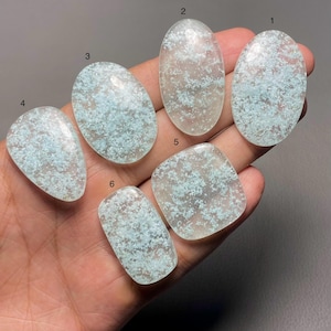 May include: Six polished gemstones in various shapes, held in a hand. The stones are translucent with a light blue, speckled pattern. Shapes include oval, teardrop, and rectangular. The gemstones are displayed against a neutral background.