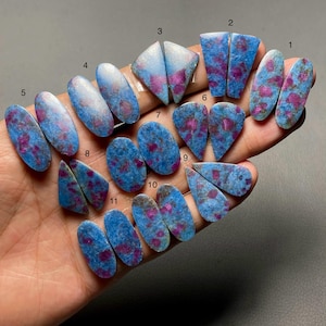 May include: A collection of eleven pairs of polished cabochon gemstones, each featuring a vibrant blue matrix with embedded magenta-colored inclusions. The stones are numbered one through eleven and display various shapes including ovals and triangles.