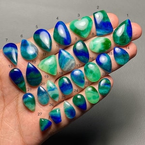 May include: A collection of 27 polished gemstones in various shapes, including teardrops, ovals, and hearts. The stones display a mix of blue and green hues, with some featuring swirling patterns. The gemstones are held in a hand, showcasing their individual colors and designs.