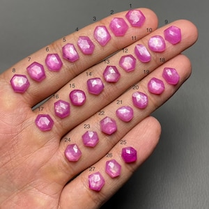 May include: Twenty-seven faceted pink gemstones of various shapes and sizes are displayed on a hand. The gemstones are numbered from 1 to 27, showcasing their polished surfaces and unique cuts. The gemstones are in a range of shapes.