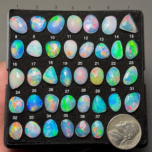 May include: A collection of 37 opals in various shapes and sizes, displayed in a black tray. The opals exhibit a range of colors, including blue, green, pink, and white. A US nickel is included for size comparison.