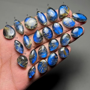 May include: A collection of polished gemstones in various shapes, including oval, teardrop, and round. The stones feature a translucent blue hue with golden, thread-like inclusions. The gemstones are numbered 1 through 23.
