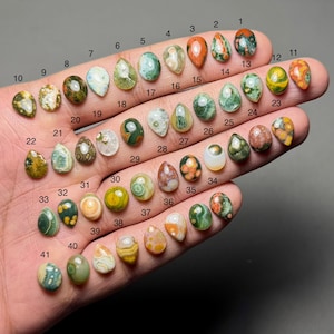 Puede incluir: Una colección de cuarenta y un cabujones de piedras preciosas pulidas en varios colores y patrones, incluyendo verde, rojo, naranja y blanco. Cada piedra está numerada y se muestra en una mano, mostrando sus formas y texturas únicas.