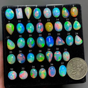 Puede incluir: Una colección de 37 piedras preciosas de ópalo expuestas en una bandeja negra. Los ópalos son de diversas formas y tamaños, con colores iridiscentes de azul, verde, rosa y amarillo.