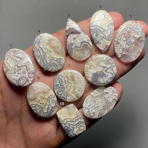Puede incluir: Diez cabujones de ágata de diferentes formas y tamaños, con intrincados patrones de blanco, gris y marrón. Los cabujones están dispuestos en semicírculo sobre una mano.