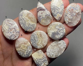 May include: Ten agate cabochons in various shapes and sizes, with intricate patterns of white, gray, and brown. The cabochons are arranged in a semi-circle on a hand.