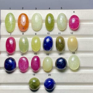 May include: A selection of twenty polished gemstones in a range of colours, such as green, orange, blue, and pink. The gemstones are oval or round, and arranged in rows. The gemstones are numbered 1 to 20.