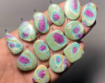 Può includere: Una collezione di tredici pietre preziose lucide di varie forme, tra cui ovali, rotonde e quadrate. Ogni pietra presenta un vivace centro magenta circondato da una matrice turchese e verde. Le pietre preziose sono esposte su una mano.