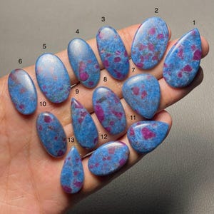 May include: Thirteen polished cabochons of blue kyanite with embedded pink ruby crystals, each numbered from one to thirteen. These smooth, oval and teardrop-shaped gemstones display a mottled texture with vibrant blue and magenta colour variations.