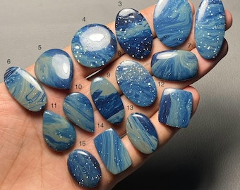 May include: A collection of fifteen polished gemstones in various shapes, including oval, teardrop, and rectangular. The stones feature a swirling blue and white pattern, some with speckled accents. The gemstones are held in a hand.