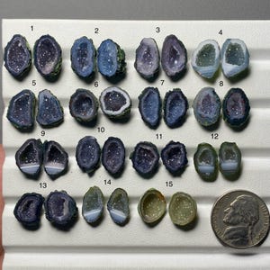 Puede incluir: Quince pares de pequeñas rodajas de geoda de ágata pulida dispuestas en filas, numeradas del 1 al 15. Las piedras presentan centros cristalinos en tonos morados, azules y verdes, algunas con bandas blancas. Se incluye una moneda de cinco centavos.